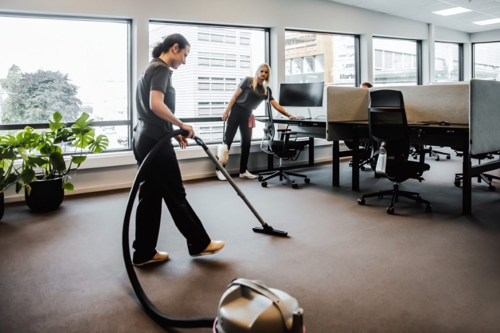 To renholdere støvsuger og tørker støv i et kontorlandskap